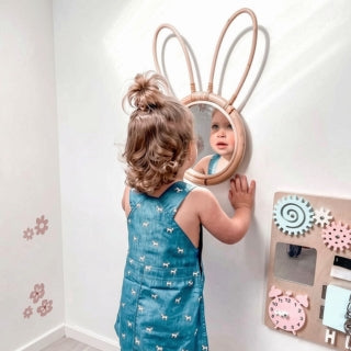 A young child with a bun hairstyle wearing a blue patterned dress looks into a round mirror with tall bunny ears on a white wall. A wooden pegboard with colorful decorative gears and a pink clock is visible on the right.