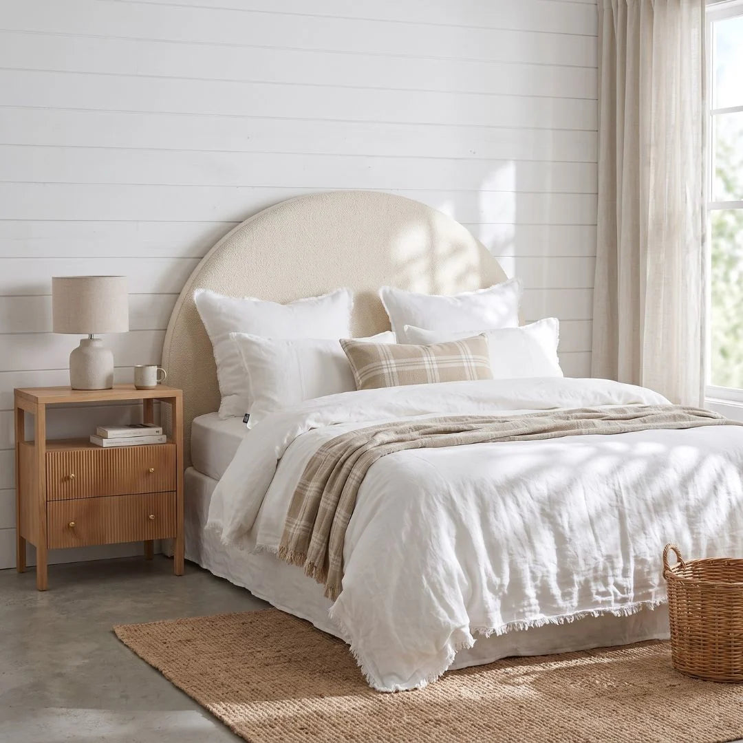 A styled bedroom scene featuring a bed dressed in white linens with a beige plaid throw blanket, set against a white shiplap wall with a light wood nightstand and window curtains.