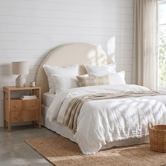 A styled bedroom scene featuring a bed dressed in white linens with a beige plaid throw blanket, set against a white shiplap wall with a light wood nightstand and window curtains.