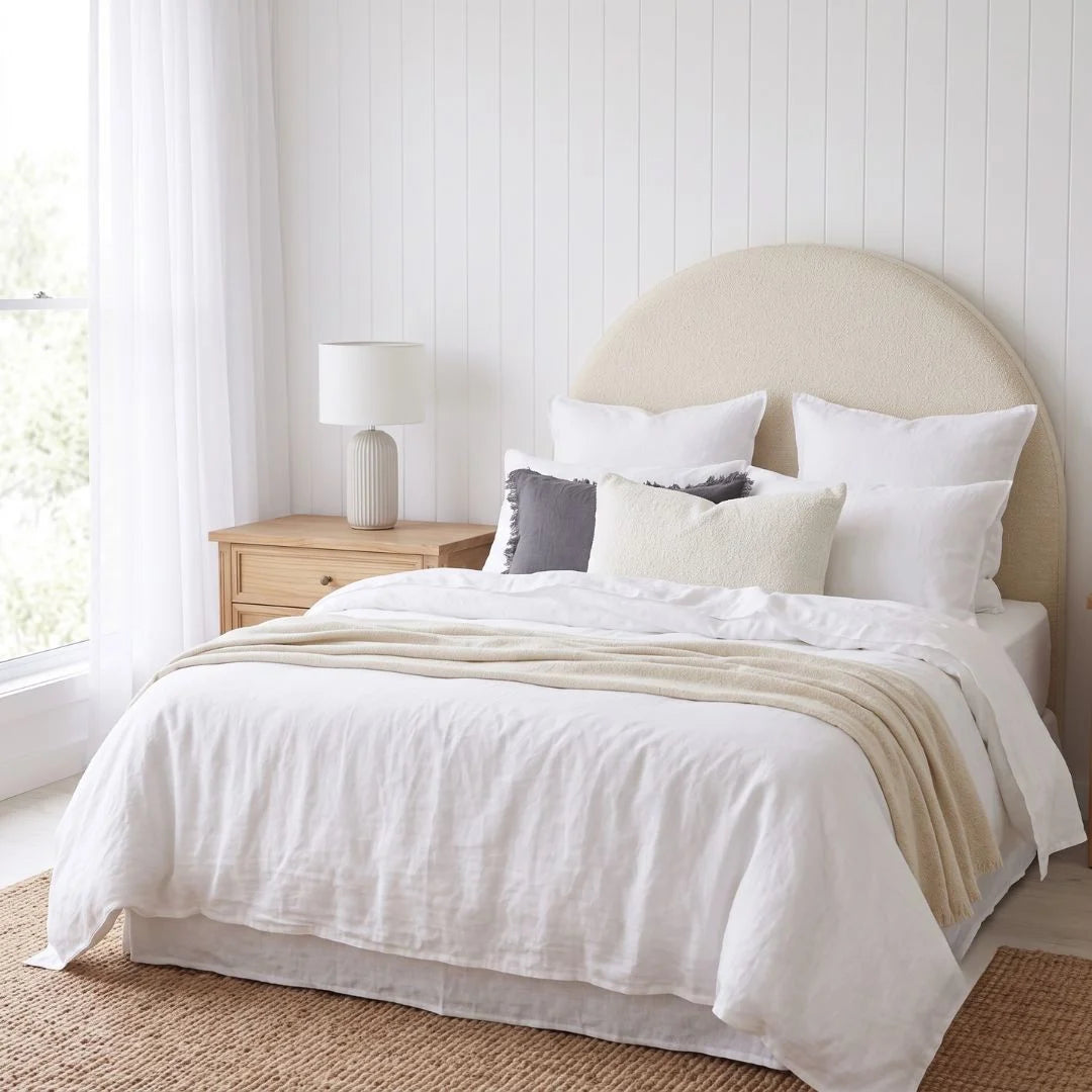 A neatly made bed in a bright bedroom with white bedding, a beige throw blanket, white pillows, a light wood nightstand, and a window with sheer curtains.