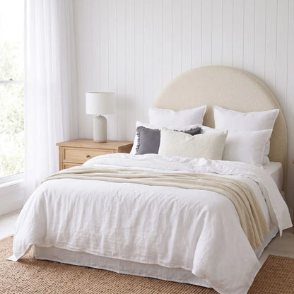 A neatly made bed in a bright bedroom with white bedding, a beige throw blanket, white pillows, a light wood nightstand, and a window with sheer curtains.