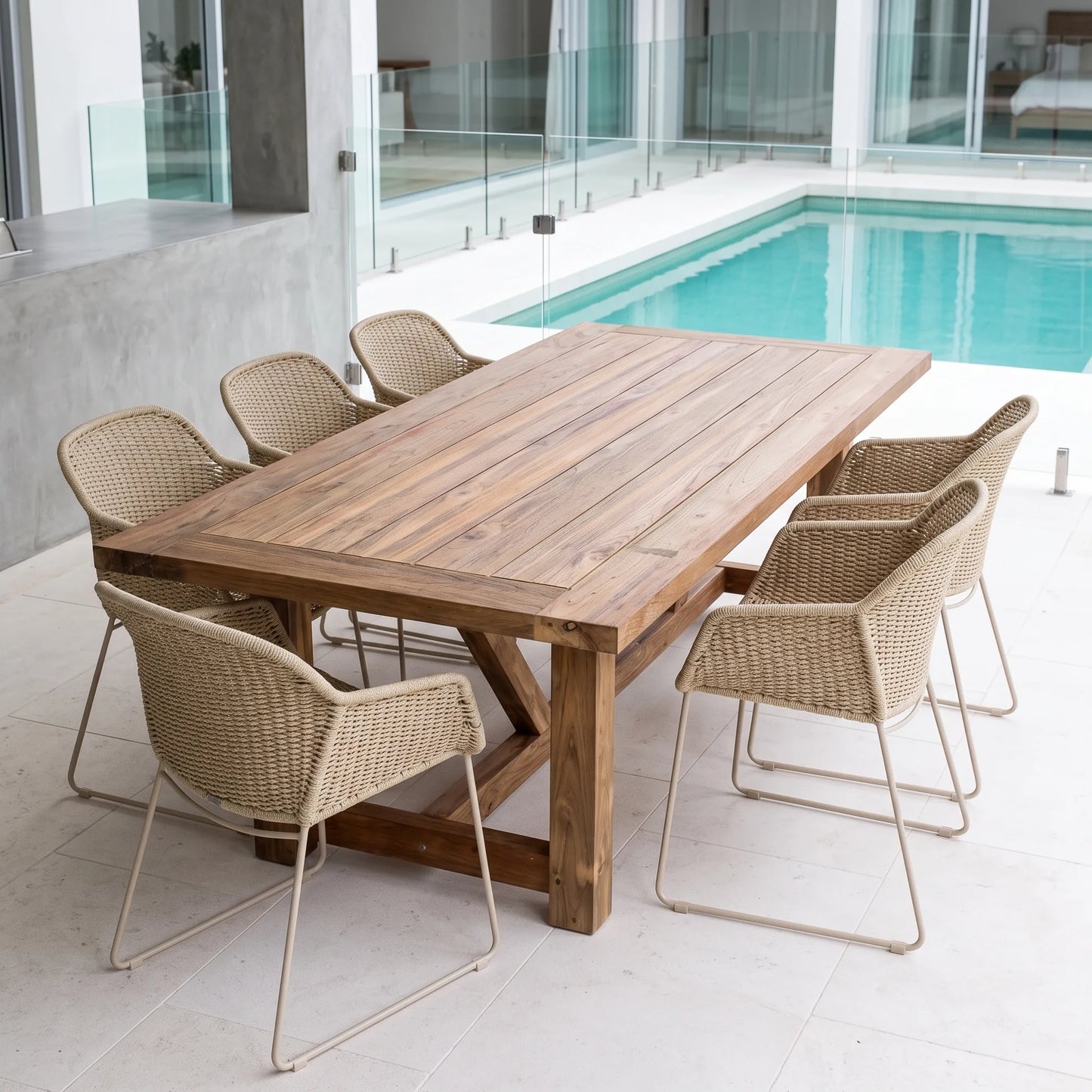 A rectangular wooden dining table with a rustic brown finish, surrounded by six woven wicker chairs with light beige metal legs, set on a tiled patio next to a swimming pool.
