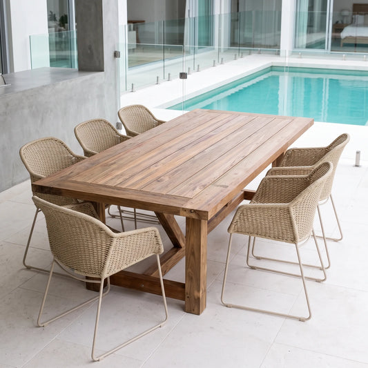 A rectangular wooden dining table with a rustic brown finish, surrounded by six woven wicker chairs with light beige metal legs, set on a tiled patio next to a swimming pool.