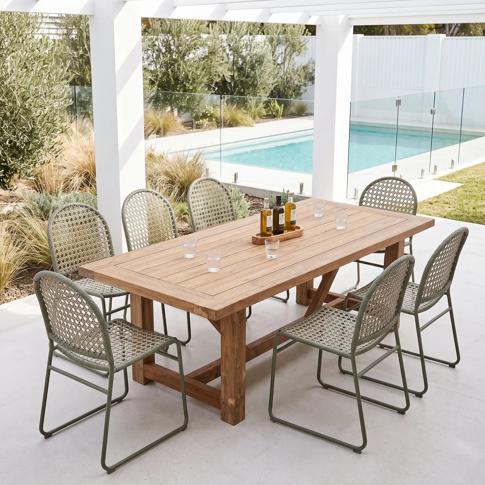 A rectangular wooden dining table with a rustic finish is set under a white pergola with six woven metal and cane chairs, overlooking a swimming pool in a backyard setting.