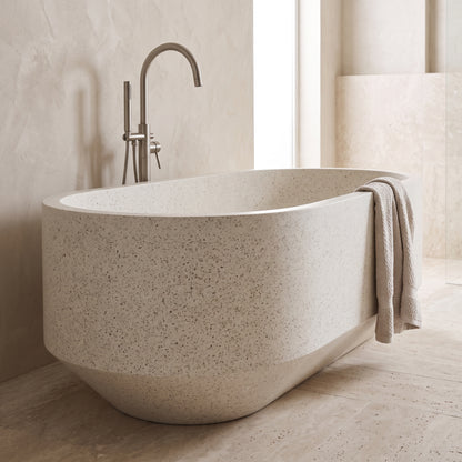 A freestanding oval bathtub in a warm beige speckled stone or concrete finish, shown in a minimalist bathroom setting with a matching beige towel draped over the side and a matte metal faucet behind it.