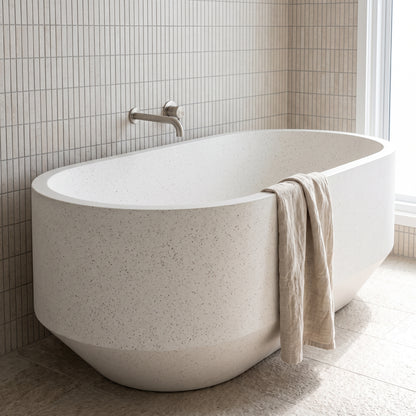 A freestanding oval bathtub in a light speckled stone or concrete finish, shown in a bathroom setting with beige wall and floor tiles, a chrome faucet, and a beige towel draped over the side.