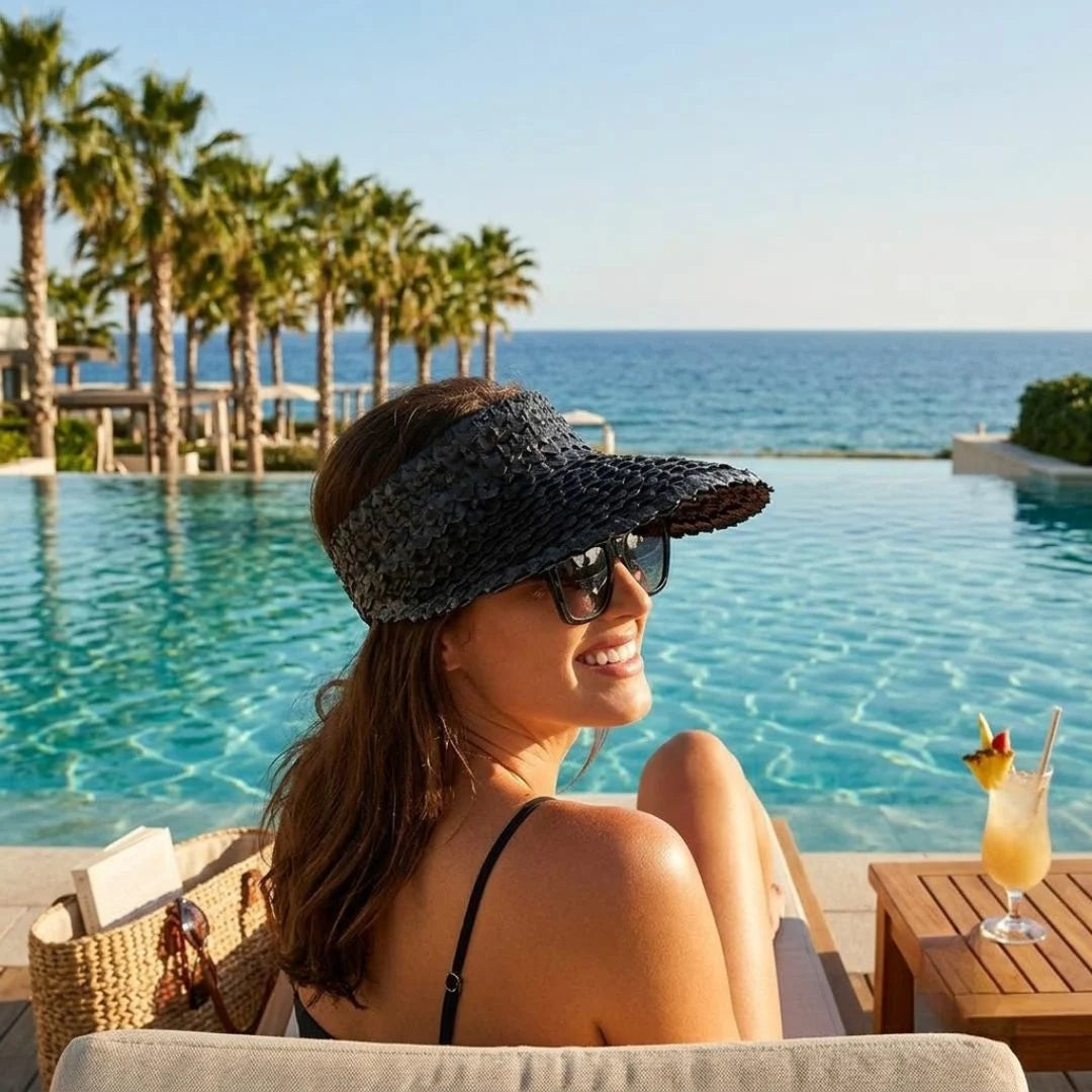 A woman wearing a black woven sun visor and sunglasses relaxes by an infinity pool overlooking the ocean, with palm trees and a cocktail on a table nearby.