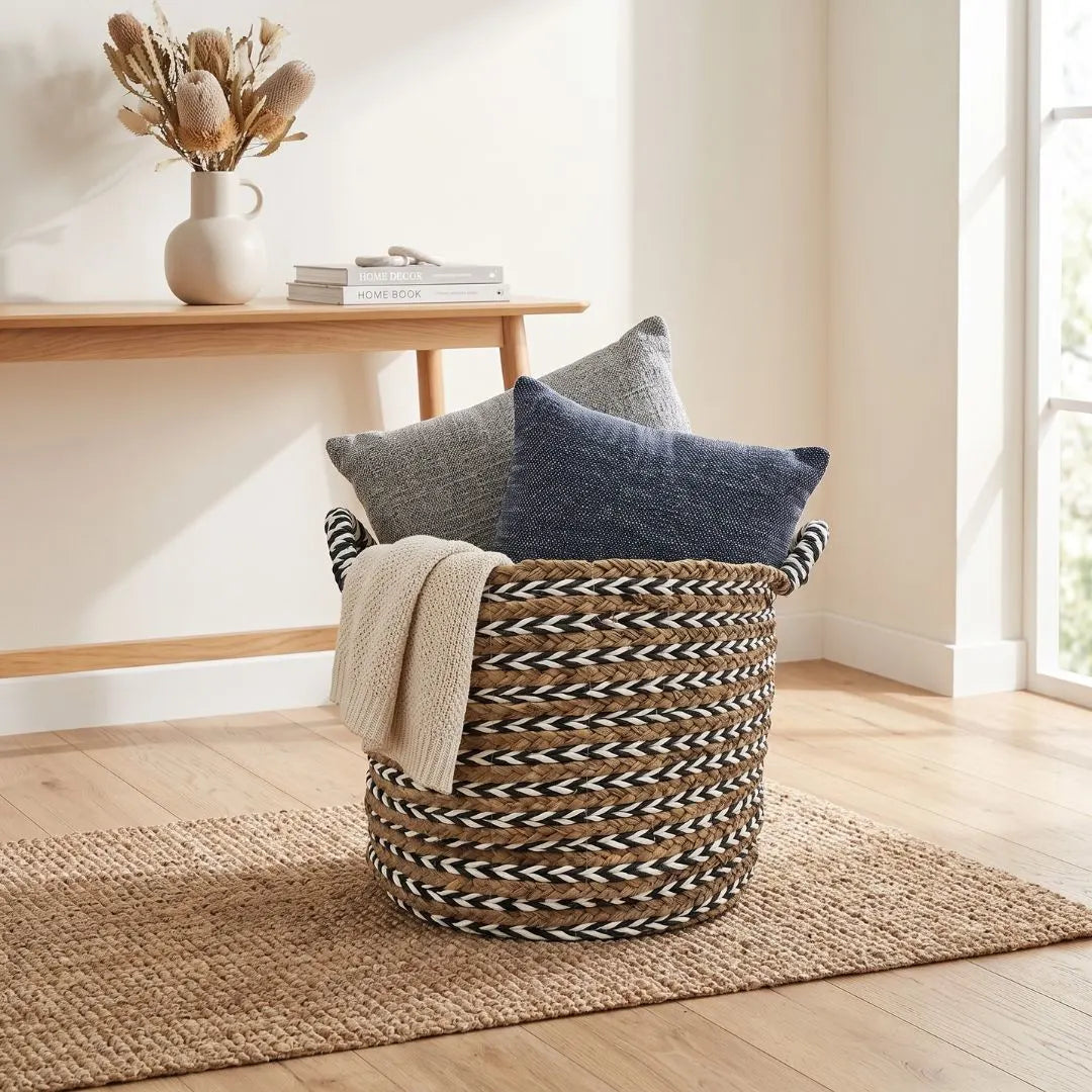 A woven round storage basket with black and beige striped handles, filled with two throw pillows and a cream knit blanket, set on a jute rug in a bright, modern room.