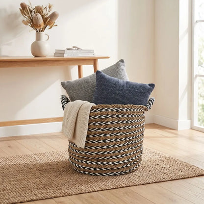 A woven round storage basket with black and beige striped handles, filled with two throw pillows and a cream knit blanket, set on a jute rug in a bright, modern room.