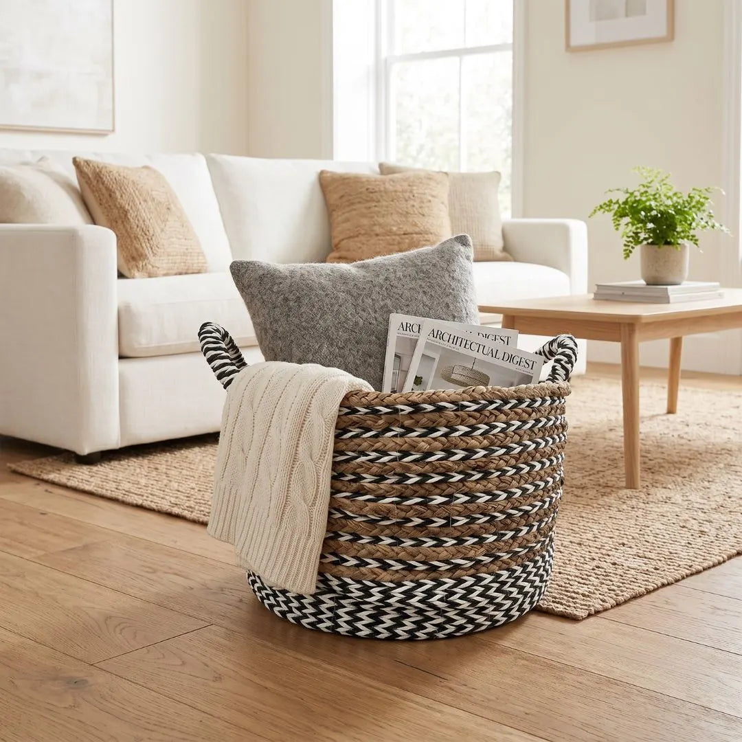 A woven storage basket with black-and-white patterned handles and a cream throw blanket draped over the side, placed in a bright living room in front of a white sofa.