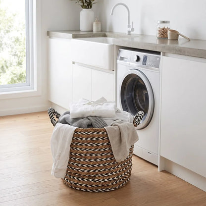 A round woven laundry basket filled with folded clothes sits on a light wood floor beside a white washing machine in a bright, modern laundry room.