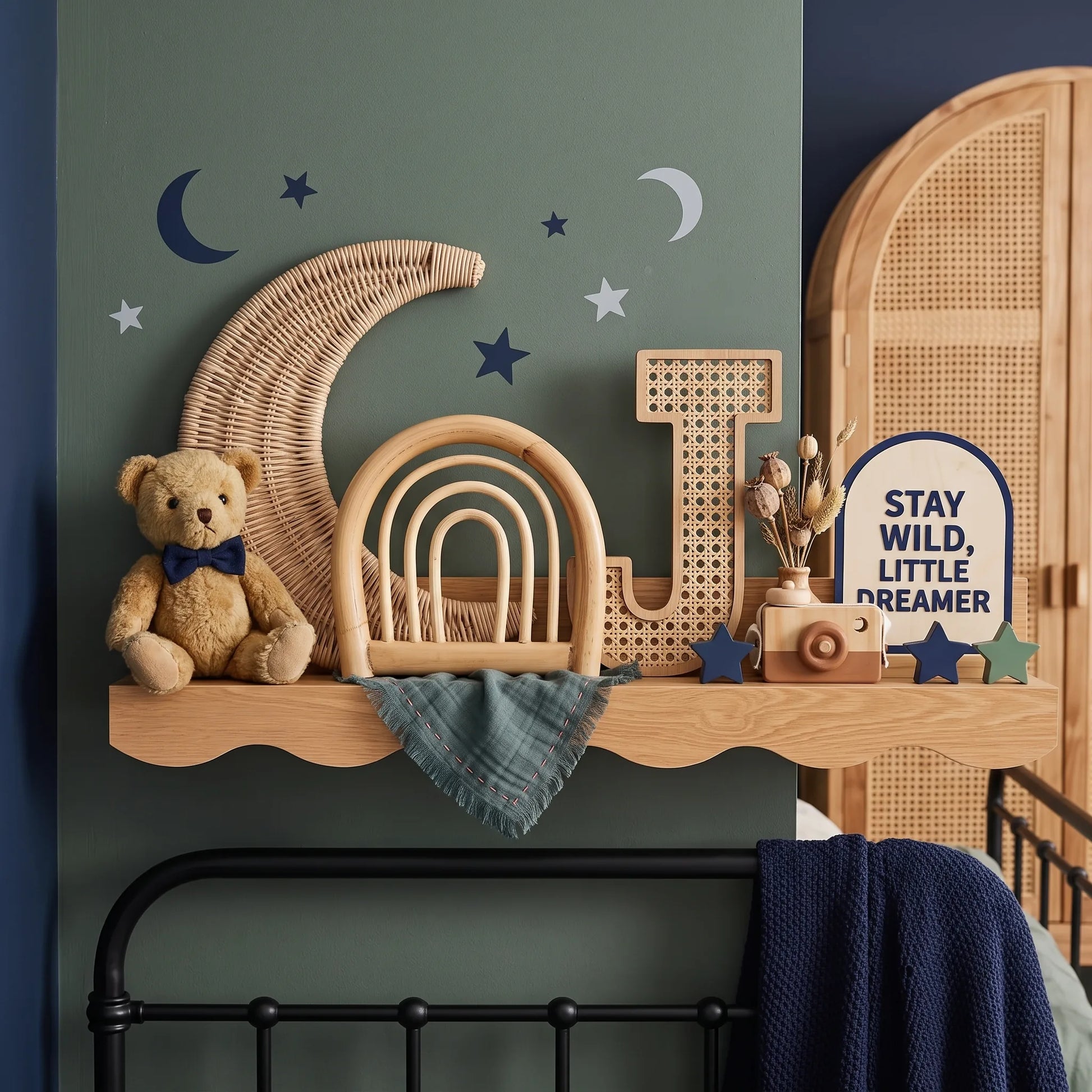 A styled nursery wall shelf with a teddy bear wearing a blue bow tie, a large wicker crescent moon, wooden letters, a small rattan chair, a toy camera, and a sign that reads 'STAY WILD, LITTLE DREAMER'. The wall is painted in a muted sage green with dark navy star and moon decals.