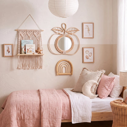 A styled bedroom corner with a wooden bed dressed in blush pink and white bedding, surrounded by boho wall decor including a macrame shelf, peach-shaped mirror, framed botanical prints, and a small arch mirror.