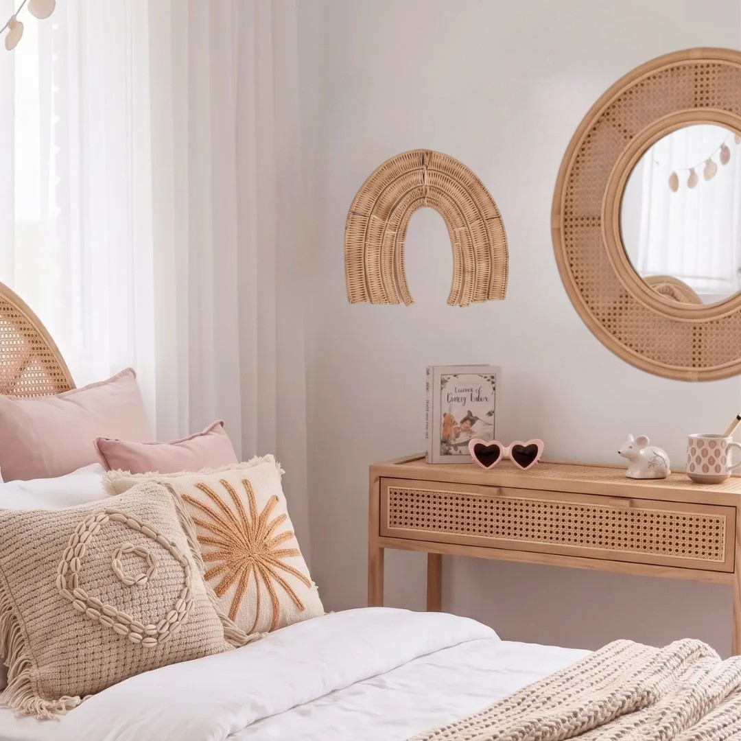 A styled bedroom corner with a bed in white bedding and pink pillows, a light wood console table, a round woven wall mirror, and a matching arch wall decor piece against a soft neutral wall.