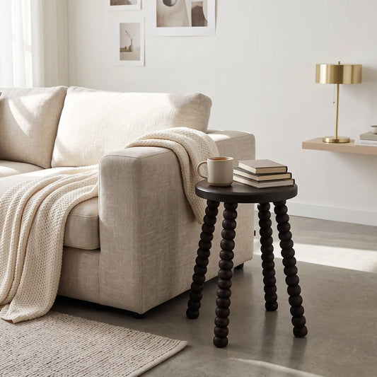 A dark round wooden side table with three knurled bead-like legs stands beside a light beige sofa in a bright living room. The table holds a mug and a stack of books, with a cream throw blanket draped over the sofa and a brass floor lamp in the background.
