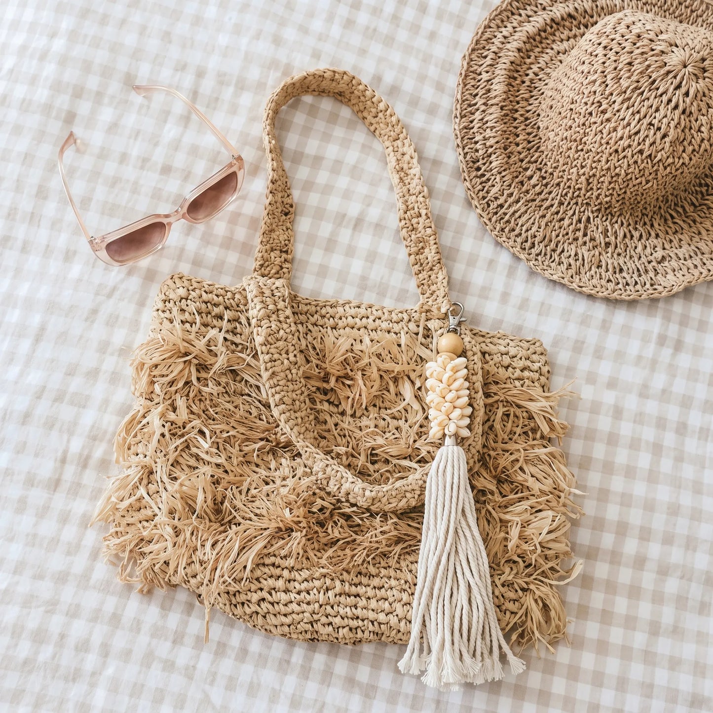A natural straw tote bag with frayed fringe detailing and a cream tassel charm, styled on a beige and white gingham fabric alongside a matching straw hat and pink sunglasses.