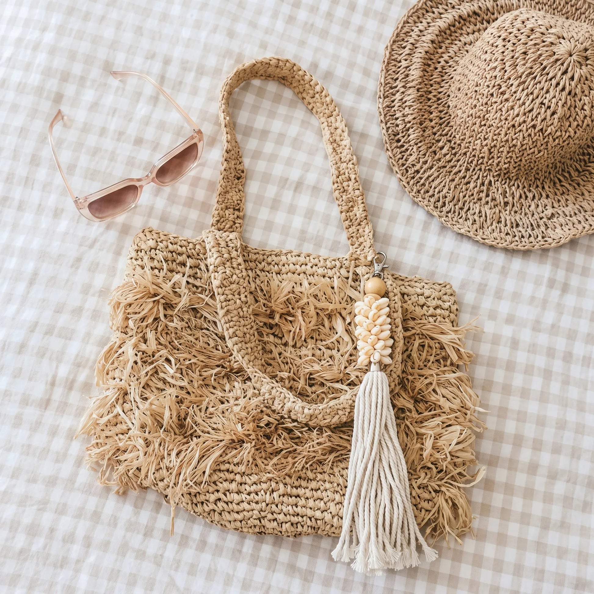 A natural straw tote bag with frayed fringe detailing and a cream tassel charm, styled on a beige and white gingham fabric alongside a matching straw hat and pink sunglasses.