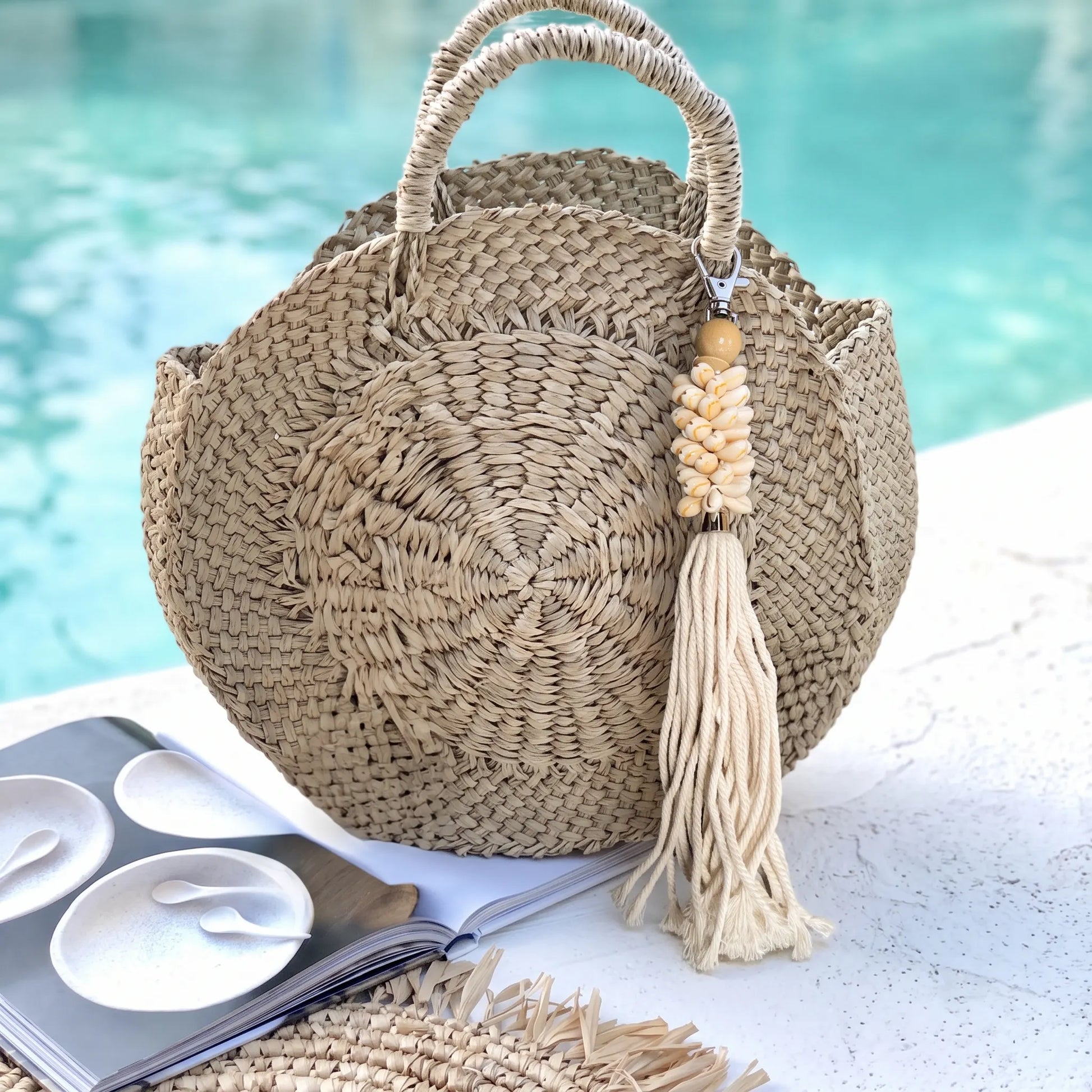 A round woven straw tote bag with two top handles and a long tassel charm, placed on a white surface near a turquoise pool. An open magazine and part of a matching woven hat are visible nearby.
