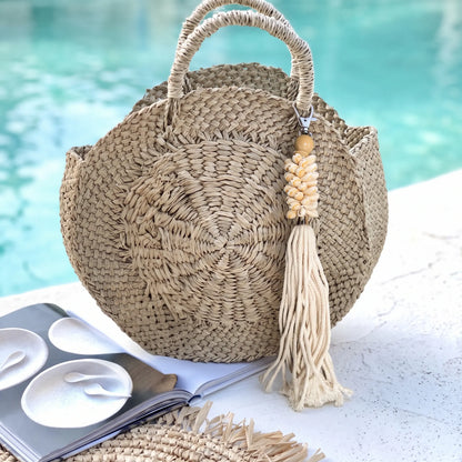 A round woven straw tote bag with two top handles and a long tassel charm, placed on a white surface near a turquoise pool. An open magazine and part of a matching woven hat are visible nearby.