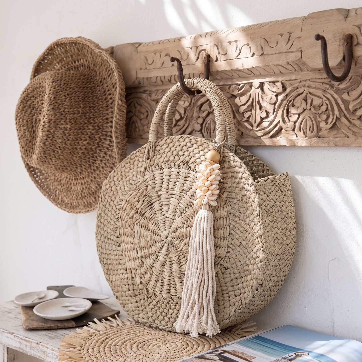 A round woven straw tote bag with dual top handles and a long cream tassel hangs from a carved wooden wall hook. A matching woven hat is draped over a carved wooden rack on the wall, with a small table and magazine below.