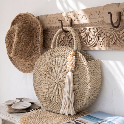 A round woven straw tote bag with dual top handles and a long cream tassel hangs from a carved wooden wall hook. A matching woven hat is draped over a carved wooden rack on the wall, with a small table and magazine below.