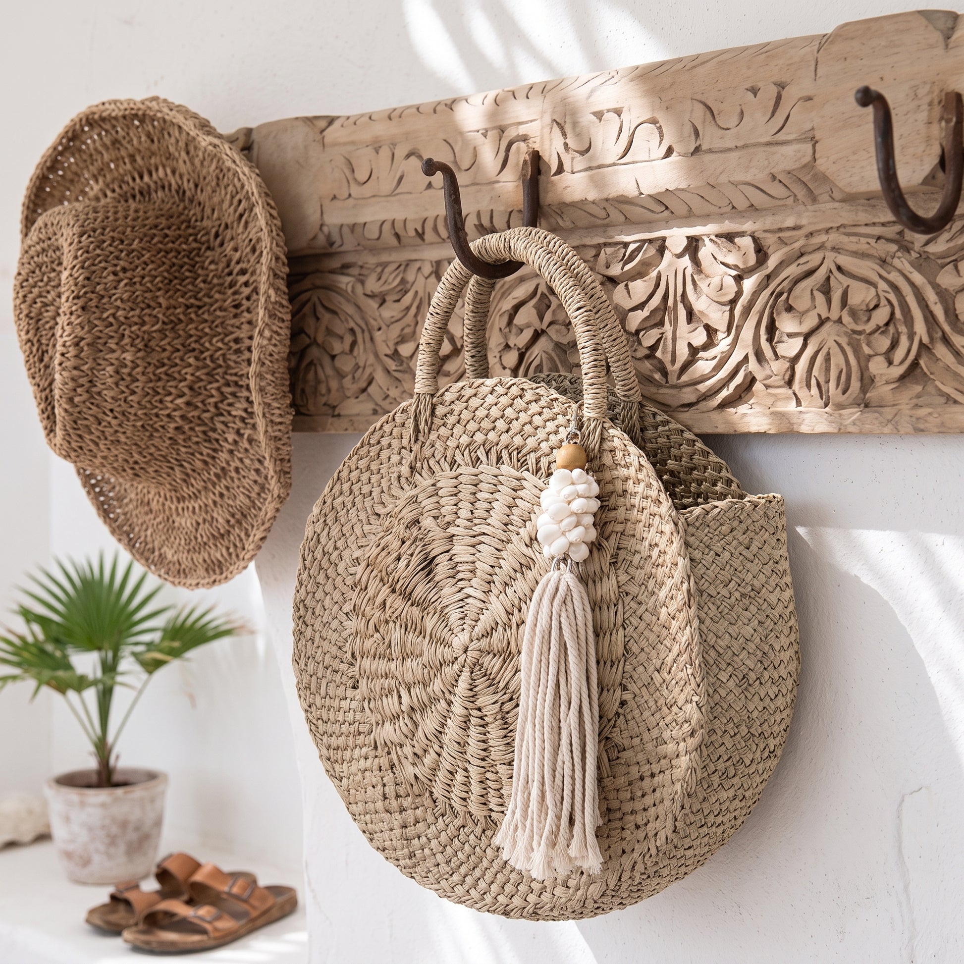 A round woven straw tote bag with double top handles and a cream tassel charm hangs from a carved wooden wall hook. A matching woven straw hat rests on the left side of the hook, with a small potted plant and sandals on a shelf below against a white wall.