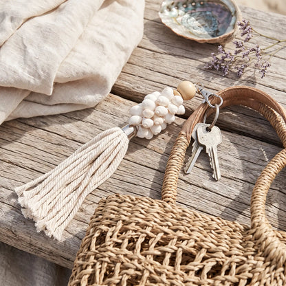 A woven straw tote bag with a cream tassel keychain resting on a rustic wooden surface, alongside a beige linen cloth, a shell, and dried flowers.