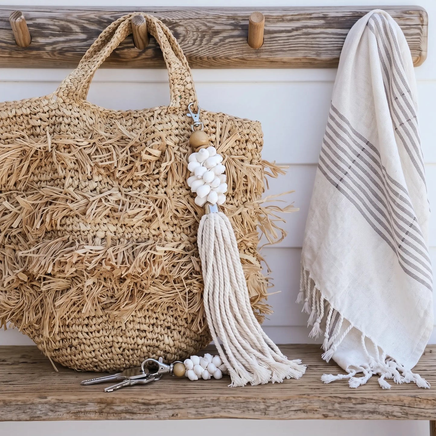 A natural woven straw tote bag with fringe and a long cream tassel charm hangs on a rustic wooden bench beside a cream striped textile towel.