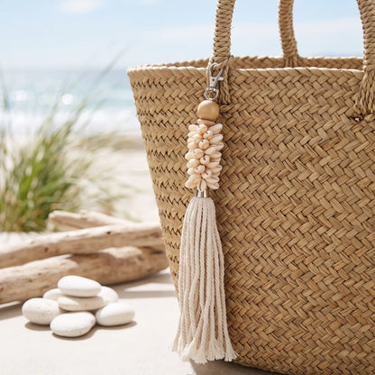 A woven straw tote bag with a cream tassel charm featuring seashells and a wooden bead, set on a sandy beach with driftwood and white stones in the background.