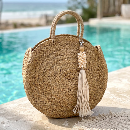 A round woven straw tote bag with dual top handles and a decorative tassel charm, photographed on a stone surface with a swimming pool and beach in the background.