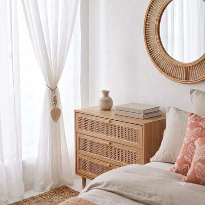 A light natural wood three-drawer nightstand with woven cane fronts, styled in a bright bedroom next to a bed with neutral linens and a pink patterned pillow. A small beige ceramic vase and books sit on top, with a round rattan wall mirror and white sheer curtains in the background.