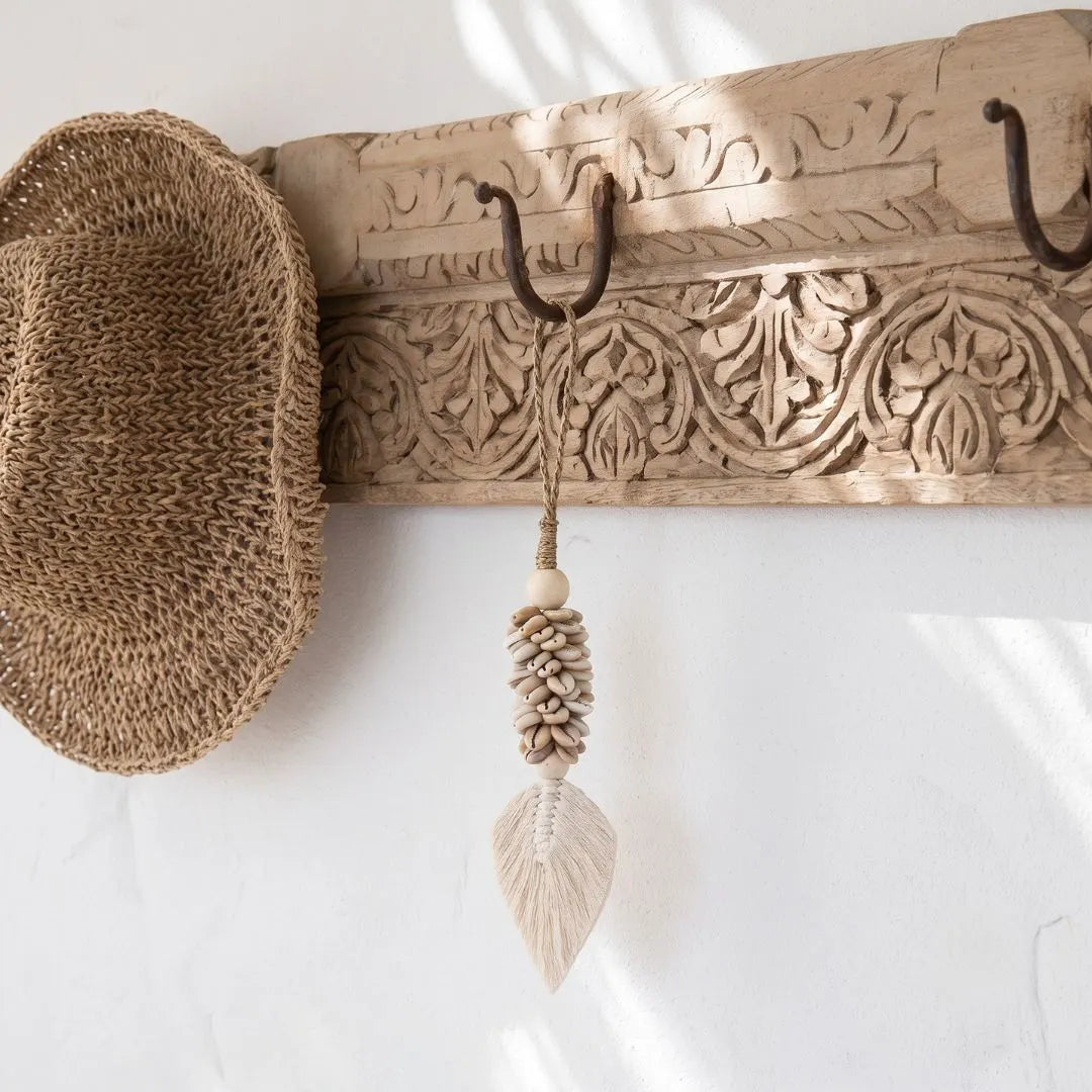 A carved wooden wall hook rack with dark metal hooks, shown mounted on a white wall. A woven straw hat hangs from the left hook, and a knotted beige decorative tassel with a leaf-shaped end hangs from the center hook.