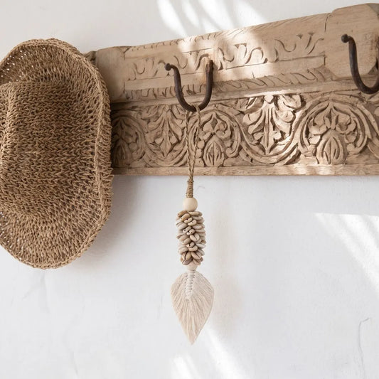 A carved wooden wall hook rack with dark metal hooks, shown mounted on a white wall. A woven straw hat hangs from the left hook, and a knotted beige decorative tassel with a leaf-shaped end hangs from the center hook.