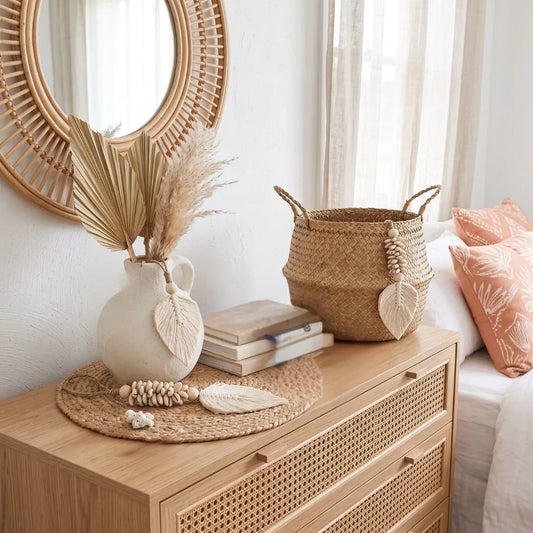 A styled bedroom scene featuring a light wood chest of drawers with woven drawer fronts, topped with a white ceramic vase holding dried palm leaves, stacked books, a round woven tray, and a natural woven storage basket beside a bed with peach pillows.
