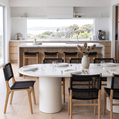 A modern dining room setup featuring a round white dining table with a thick cylindrical pedestal base, surrounded by black woven-back wooden chairs with light wood frames. The scene is set in a bright, contemporary kitchen with a large window overlooking water and trees.