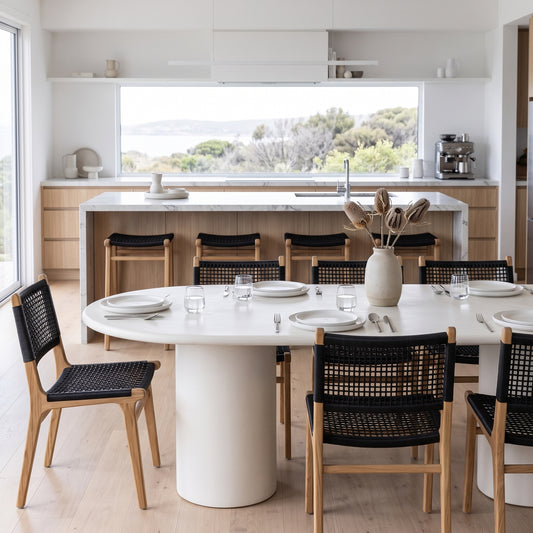 A modern dining room setup featuring a round white dining table with a thick cylindrical pedestal base, surrounded by black woven-back wooden chairs with light wood frames. The scene is set in a bright, contemporary kitchen with a large window overlooking water and trees.