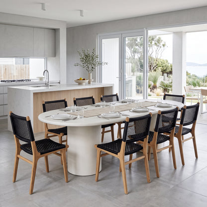 A modern dining room scene with a round white dining table surrounded by eight wooden chairs with black woven seats, set in a bright contemporary kitchen with large windows and outdoor views.