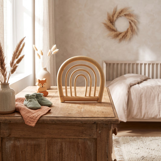 A light natural wood baby tooth holder with a rainbow arch design sits on a rustic wooden chest in a warm, sunlit nursery. The scene is styled with a ceramic vase of pampas grass, sage green baby booties, a small wooden fox toy, and a neutral bed in the background.