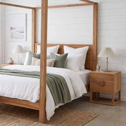 A light natural wood four-poster bed with white bedding, sage green pillows, and a sage green throw blanket, styled in a bright, modern bedroom with white shiplap walls, matching wood nightstands, and lamps.