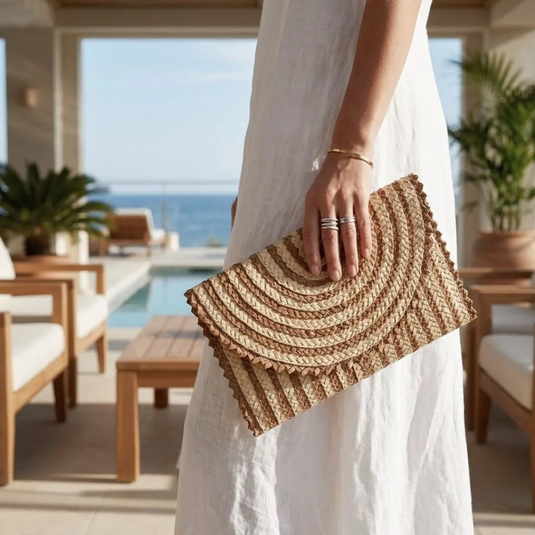 A woman in a white dress holds a woven straw clutch bag with a scalloped edge and concentric circular texture, set against a sunny outdoor patio background with a pool and ocean view.