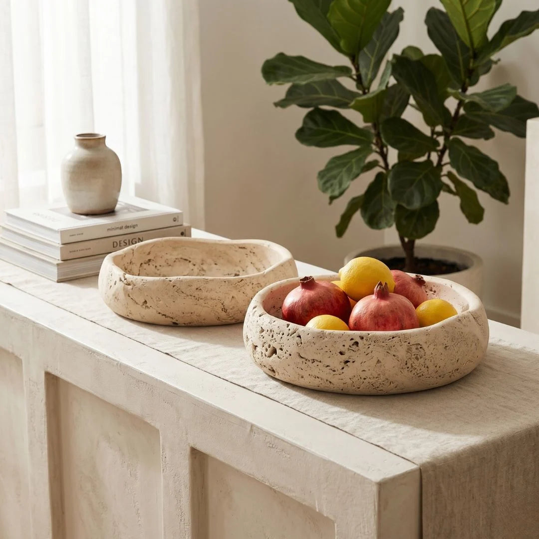 Two travertine stone bowls on a wooden surface with fruits and a plant in the background.