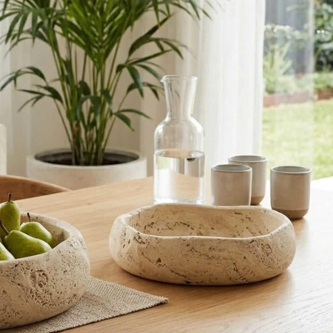Decorative travertine stone bowls and ceramic cups on a wooden table with a plant in the background.