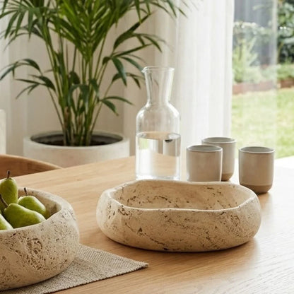 Decorative travertine stone bowls and ceramic cups on a wooden table with a plant in the background.