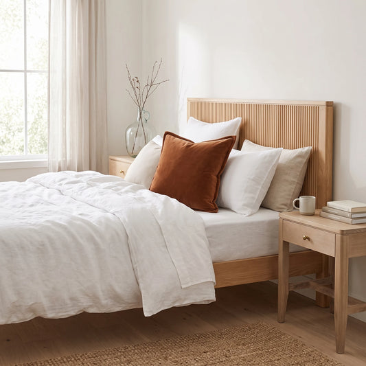 A light natural wood bed frame with a slatted headboard, styled with white bedding and a rust-orange accent pillow in a bright, modern bedroom.
