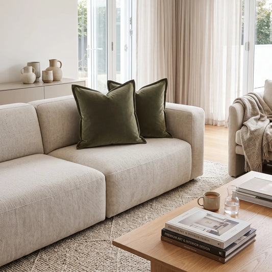 A modern light beige fabric sofa in a bright, contemporary living room, styled with two dark olive green velvet throw pillows. A wooden coffee table with books and a mug sits in the foreground, and a neutral armchair is visible on the right.
