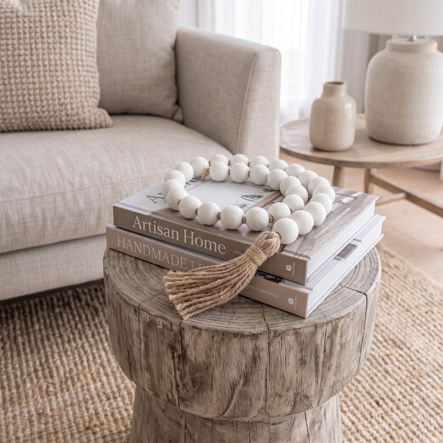 A decorative strand of matte white wooden beads with a natural jute tassel draped over stacked books on a rustic round wood side table in a softly styled living room.
