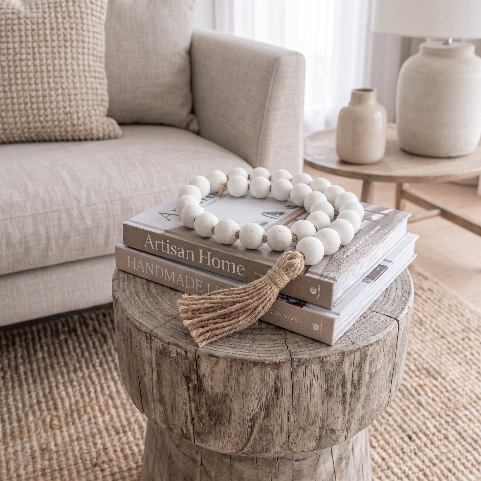 A decorative strand of matte white wooden beads with a natural jute tassel draped over stacked books on a rustic round wood side table in a softly styled living room.