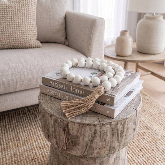 A decorative strand of matte white wooden beads with a natural jute tassel draped over stacked books on a rustic round wood side table in a softly styled living room.