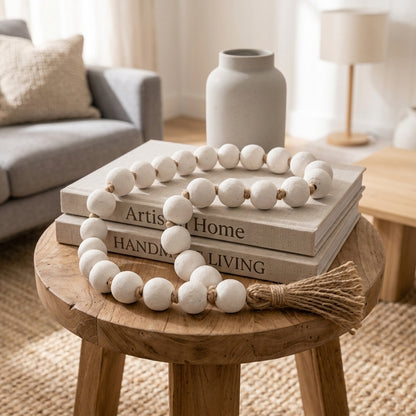 A decorative garland made of matte white round beads with small natural wood spacers and a jute tassel, draped over stacked books on a wooden stool in a softly styled living room.