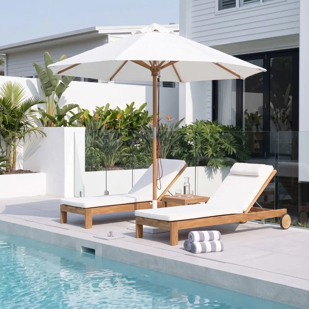A white patio umbrella with a wooden pole stands beside two wooden lounge chairs with white cushions on a poolside deck. White walls, plants, and a modern house are visible in the background.